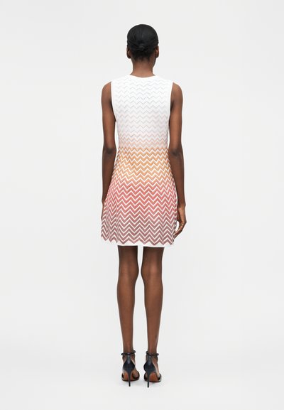 Γυναίκα στέκεται με την πλάτη στην κάμερα, φορώντας αμάνικο φόρεμα με σχέδιο ζιγκ-ζαγκ που ξεθωριάζει από λευκό σε πορτοκαλί και κόκκινο, με μαύρες ψηλοτάκουνες γόβες.