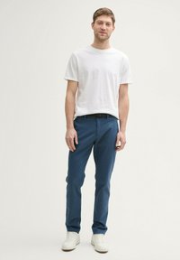 Junger Mann steht mit einem schlichten weißen T-Shirt, blauen Hosen mit einem schwarzen Gürtel und weißen Sneakers vor einem neutralen Hintergrund.