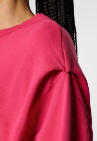 Sweatshirt rosa com uma textura suave, decote redondo e detalhe franzido na costura do ombro. Corte casual e design versátil.