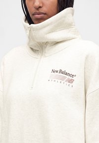 Άτομο που φοράει ένα ανοιχτό μπεζ πουλόβερ New Balance Athletics με ψηλό γιακά με φερμουάρ και λογότυπο σε καφέ χρώμα στο αριστερό στήθος.
