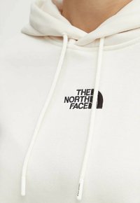Biała polarowa bluza z kapturem z przednią kieszenią, sznurkami oraz czarnym haftowanym logo "The North Face" po lewej stronie klatki piersiowej.