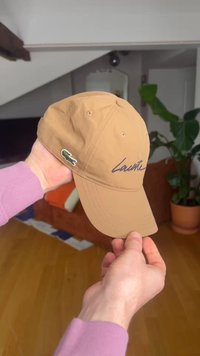 Mâini care țin o șapcă baseball Lacoste de culoare bronz, cu logo brodat și emblema crocodilului, într-o cameră de interior cu canapea și plantă în fundal.