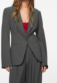 Blazer gris à la silhouette ajustée, col à revers net, fermeture par un bouton unique et manches longues. Porté sur un top rouge avec des boutons.