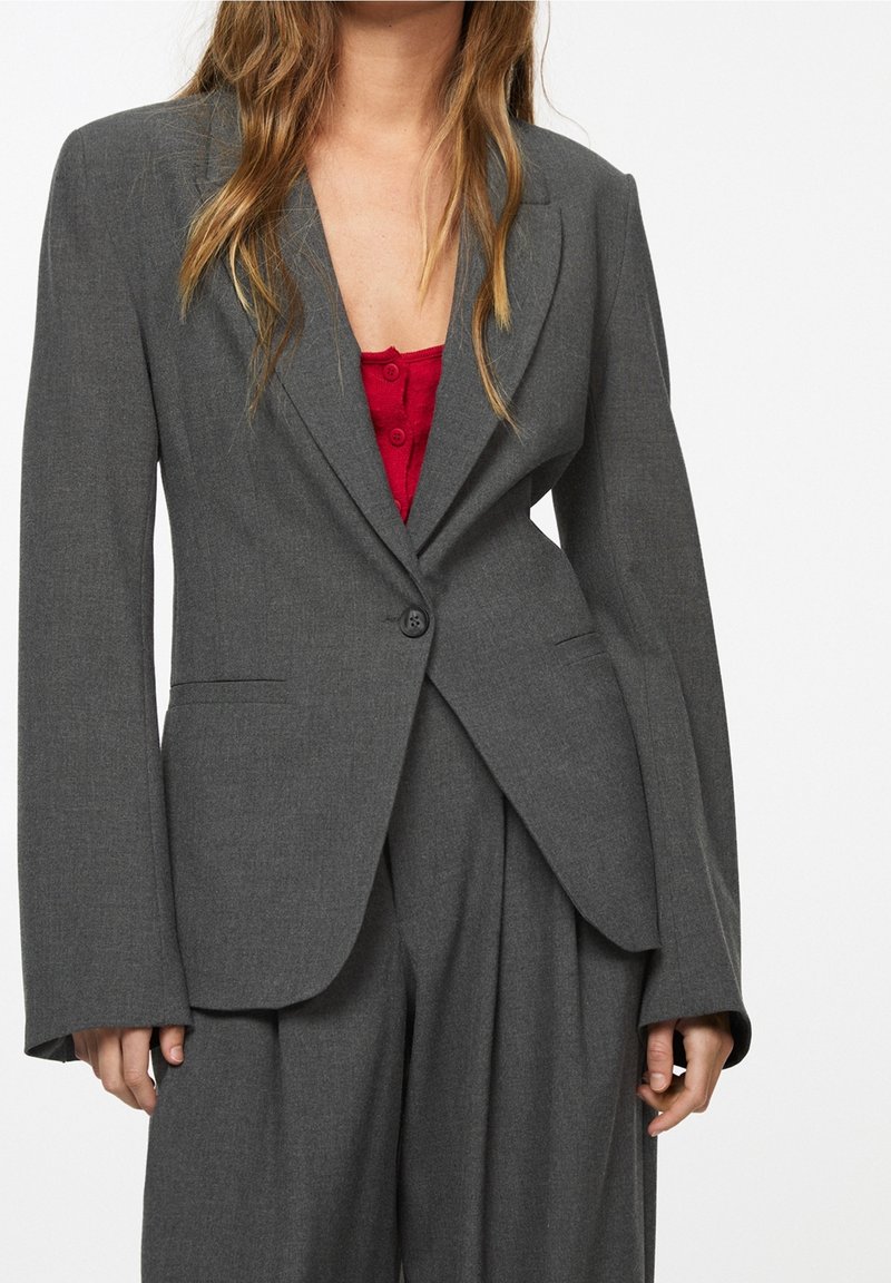 Blazer gris à la silhouette ajustée, col à revers net, fermeture par un bouton unique et manches longues. Porté sur un top rouge avec des boutons.