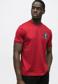 Rød bomuld T-shirt med korte ærmer, rund halsudskæring og sort diamantformet logo på venstre bryst. Glat tekstur, enkelt design.