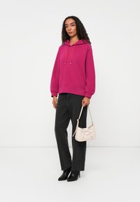 Magenta Kapuzenpullover mit Kordelzug, schwarze weit geschnittene Jeans und spitze schwarze Absätze. Hält eine kleine cremefarbene Handtasche mit Schleifendetail.