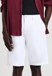 White shorts made from a lightweight fabric, featuring a button closure and side pockets. The garment has a clean, tailored shape.