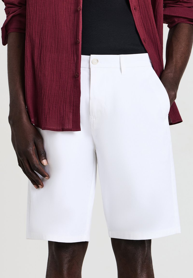 White shorts made from a lightweight fabric, featuring a button closure and side pockets. The garment has a clean, tailored shape.