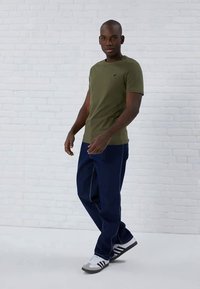 Man wearing olive green t-shirt, dark blue jeans, and gray-and-white sneakers stands against white brick wall.