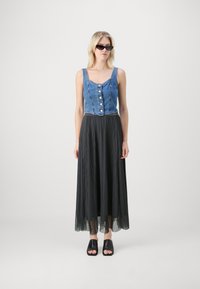 Denim kratka majica z gumbom, kombinirana s črno plisirano maksi krilo in črnimi mule čevlji z odprtimi prsti. Sončna očala dopolnjujejo videz.