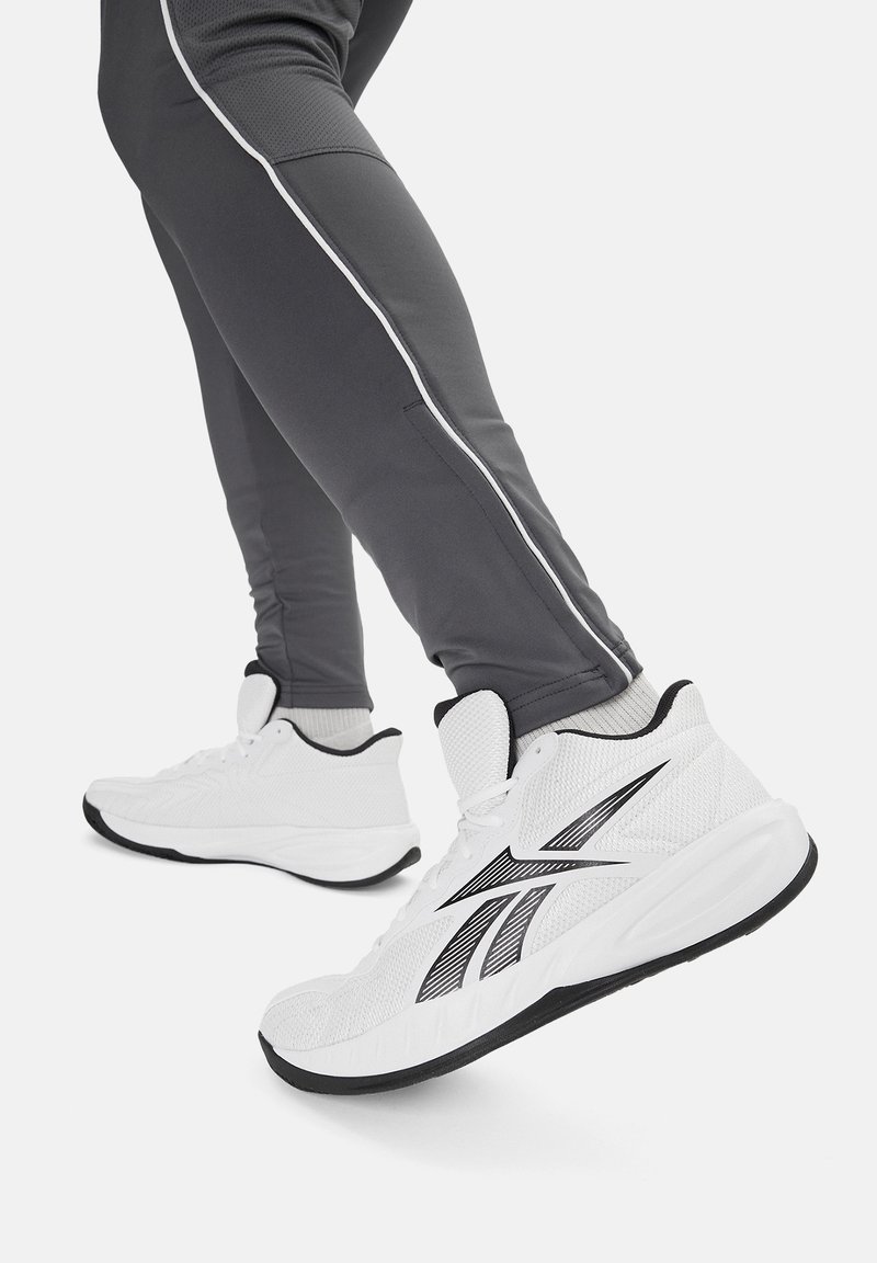 Weiße Sportschuhe mit Mesh-Obermaterial, schwarzen Akzenten und einer strukturierten Sohle, kombiniert mit grauen Sporthosen mit weißen Seitenstreifen.