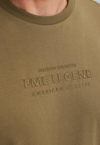 Olivenfarbendes Baumwoll-T-Shirt mit Rundhalsausschnitt und eingeprägtem Text "FREEDOM UNLIMITED PME LEGEND AMERICAN CLASSIC". Weiche Textur.