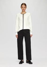 Witte blouse met lange mouwen, zwarte accenten en een keyhole-halslijn, gecombineerd met zwarte wijde broek en glanzende zwarte schoenen.