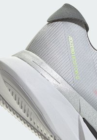 Grauer Sportschuh mit einem strukturierten Mesh-Obermaterial, reflektierenden Akzenten und einer weißen Zwischensohle. Verfügt über einen gepolsterten Schuhkragen und eine gemusterte Laufsohle.