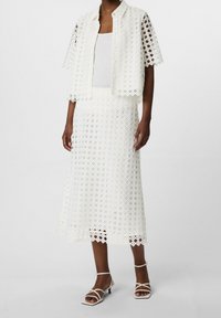 Ensemble en dentelle blanche comprenant une veste courte à motif losange, accompagnée d'une jupe mi-longue assortie et de sandales blanches à lanières.