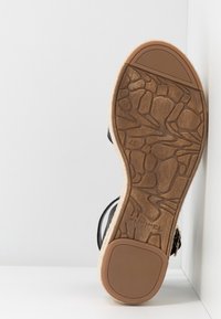 Sandalsula med texturerad stenmönster, svarta remmar och beige kanter lutande mot vit hörnyta.