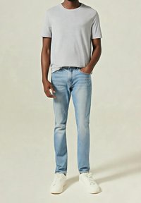 Homme portant une chemise à manches courtes gris clair, un jean bleu clair, des baskets blanches, debout avec une main dans la poche devant un fond uni.