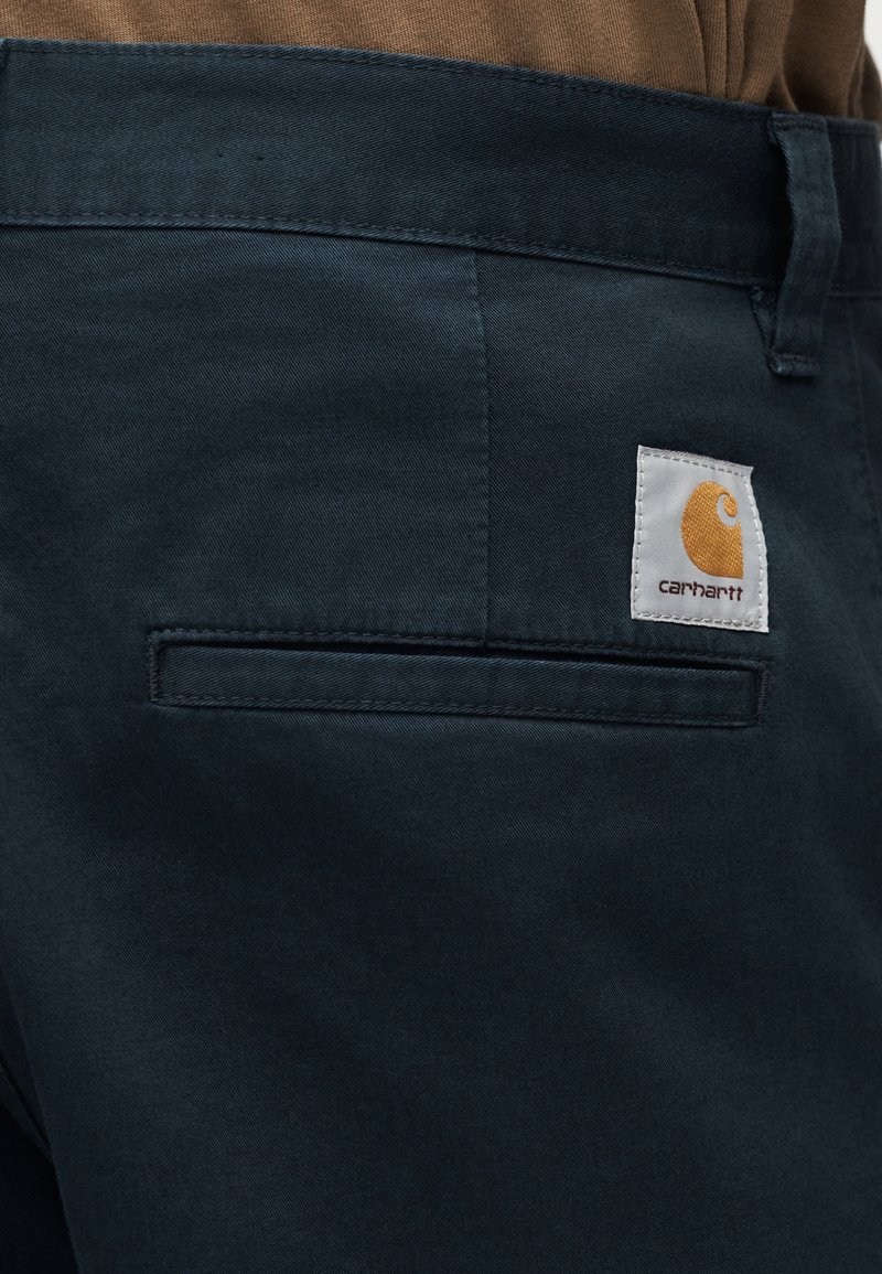Nahaufnahme der Gesäßtasche einer marineblauen Hose mit Carhartt-Logo-Patch und einem Teil eines braunen Hemds, das am Bund sichtbar ist.
