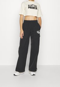 Camiseta recortada de color crema con el logotipo negro de "NIKE", combinada con pantalones cargo holgados negros que tienen dos bolsillos laterales y un logotipo blanco.