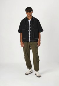 Black oversized button-up shirt with white stitching, paired with olive cargo pants featuring multiple pockets and white sneakers.