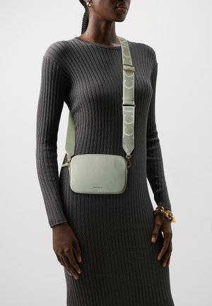 Lichtgroene leren schoudertas met een gestructureerde afwerking, gouden hardware en een gestreepte verstelbare schouderband. Gedragen over een donkere geribbelde jurk.