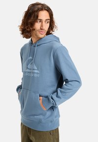 Modrá mikina s kapucí vyrobená z měkké tkaniny, s přední kapsou a logem na hrudi. Navržena s kapucí na stažení a žebrovanými manžetami.