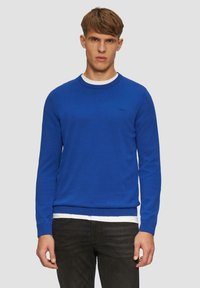 Pull bleu à col rond avec des poignets et un ourlet côtelés, superposé sur un T-shirt blanc, associé à un jean foncé. Texture de tissu tricoté douce.
