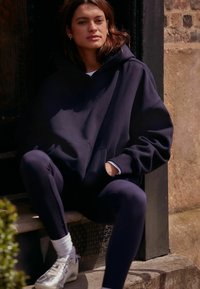 Marinblå oversized hoodie med framfickor, kombinerad med marinblå leggings och silverfärgade träningsskor, sittande på ett steg mot en strukturerad vägg.