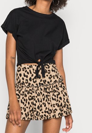 Kobieta ubrana w czarny krótki top związywany z przodu na supeł oraz beżową mini spódniczkę w panterkę z falbankami, stojąca na jednolitym tle.