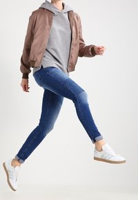 Lichtbruine bomberjack over een grijze hoodie, blauwe skinny jeans en witte sneakers met groene accenten, wat een casual, gelaagde look laat zien.