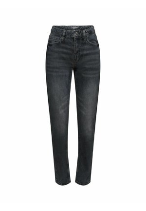 Personne portant un jean noir délavé taille haute avec une ceinture lilas, un haut jaune à manches longues et des baskets noires et beige, debout avec une main dans la poche.