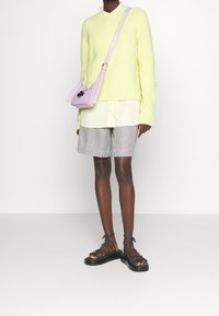 Leuchtend gelber Strickpullover, weißes Hemd mit Kragen, gestreifte Shorts und grüne Sandalen mit Riemchen. Rosa Umhängetasche mit Metallschmuck.