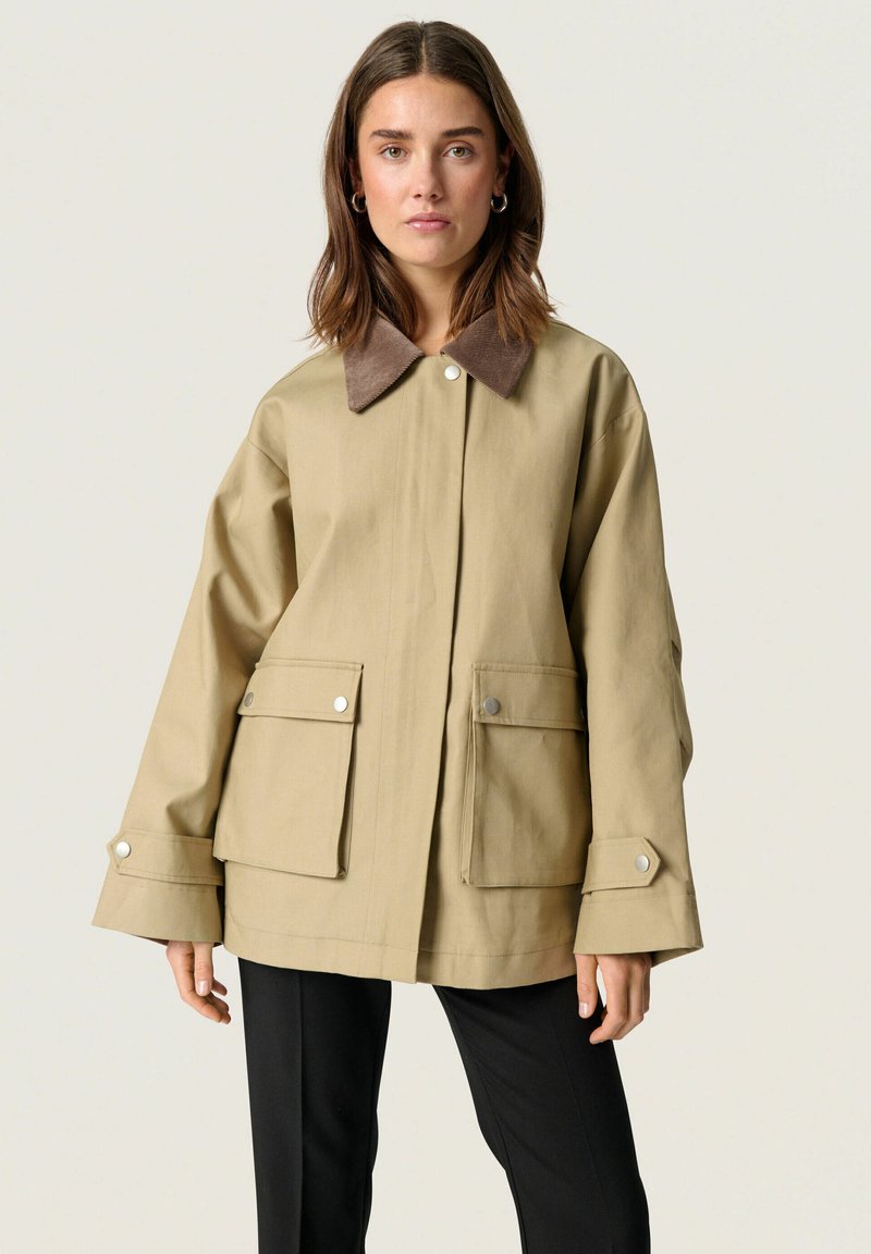 Veste oversized beige avec un col en velours côtelé marron, de grandes poches avant, des boutons-pression et des manches larges. Portée par-dessus un pantalon noir.