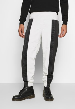 Pantalons de sport blancs et noirs avec une bande latérale noire ornée d'un logo, une taille élastique et des chevilles resserrées. Associés à des bottes noires.