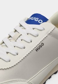 Biały sneaker HUGO z białymi sznurowadłami, siateczkową teksturą oraz niebieską metką na języku z nazwą marki "HUGO" widoczną z boku.