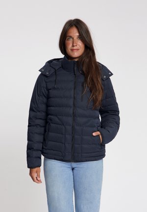 Vrouw met lang bruin haar, gekleed in een marineblauwe doorgestikte jas met ritssluitingen en lichtblauwe spijkerbroek, staand tegen een effen achtergrond.