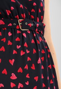 Robe marine avec un motif en forme de cœur en rouge vif, dotée d'une ceinture assortie avec une boucle dorée, fabriquée à partir d'un tissu lisse.