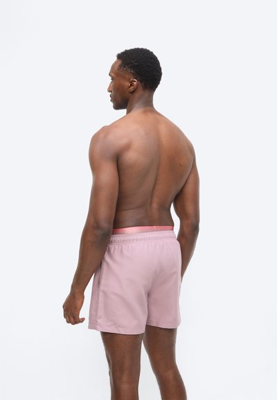 Άνδρας με σκουρόχρωμο δέρμα και κοντά μαλλιά φοράει ανοιχτό ροζ μαγιό, στέκεται με γυμνή πλάτη και κοιτάζει προς τα αριστερά σε λευκό απλό φόντο.