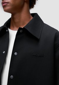 Sort jakke med krave, sølv snaps og prægede "WRSTBHVR" logo. Glat stof med mat finish.