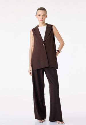 Kvinde med tilbagelagt hår iført mørkebrunt ærmeløst blazer og bukser med brede ben, hvid top og sandaler mod ensfarvet hvid baggrund.