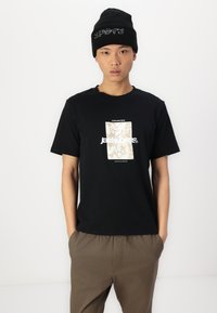 Sort bomulds T-shirt med et cremefarvet grafisk rektangel og logo i hvid. Båret sammen med lysebrune, løstsiddende bukser og en sort strikket beanie.
