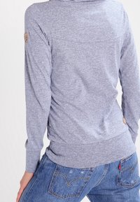 Grau hooded Sweatshirt mit langen Ärmeln, geripptem Saum und Logodetail am Ärmel. Getragen mit blauen Jeans. Weiche Stofftextur.