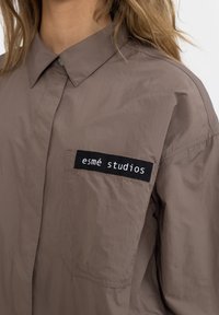 Hnědá košile s knoflíky a černou nášivkou na kapse s nápisem "esmé studios", kterou nosí osoba s dlouhými blond vlasy.