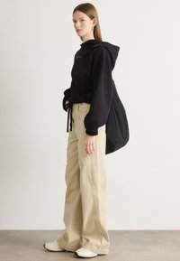 Felpa nera con cordini regolabili, pantaloni beige chiaro a gamba larga e scarpe da ginnastica bianche. Borsa nera appesa a una spalla. Texture lisce.