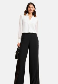 Blusa bianca con colletto e dettagli dei bottoni, abbinata a pantaloni neri a gamba larga a vita alta. Una borsa nera completa il look.