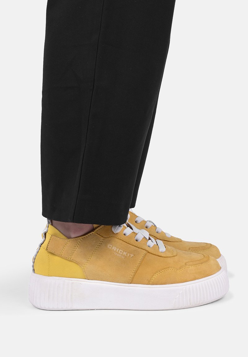 Person trägt senfgelbe Wildleder-Sneaker mit weißen Schnürsenkeln und dicker weißer Sohle, kombiniert mit schwarzen Hosen.