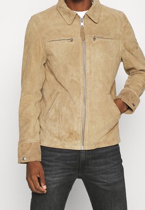 Homme portant une veste zippée en daim beige avec deux poches zippées poitrine et des poches latérales, associée à un jean gris foncé.