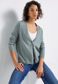 Cardigan vert clair avec un col en V et un fermoir à boutons, présentant des poignets et un ourlet côtelés, porté sur un haut blanc et assorti à un jean bleu.