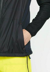 Schwarze Steppweste aus strukturiertem Stoff, mit Reißverschluss vorne und seitlichen Taschen. Akzentuiert mit orangefarbenen Fäden nahe dem Saum und den Ärmeln.