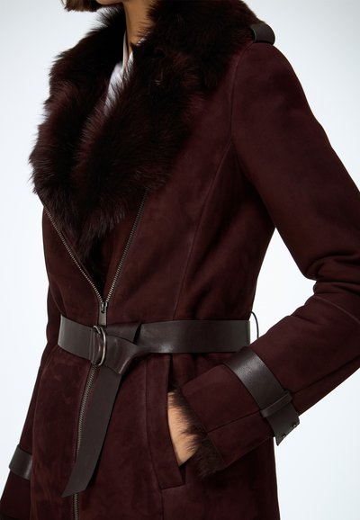 Burgundský semišový kabát s kožušinovým golierom, opaskom v páse a koženými detailmi na rukávoch. Má predný zips a hladkú textúru.